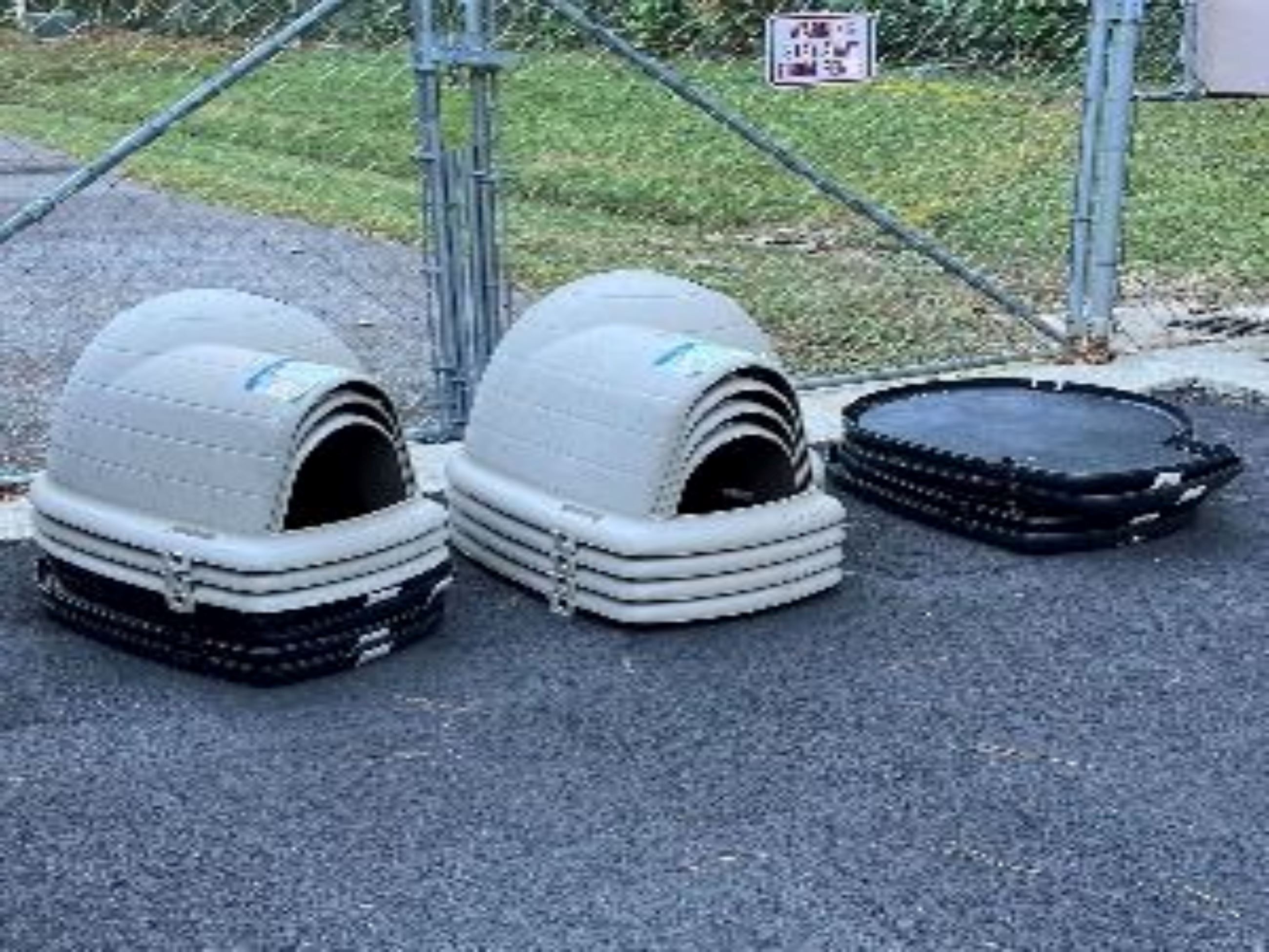 Dog houses stacked up on pavement in front of a chain link fence. 